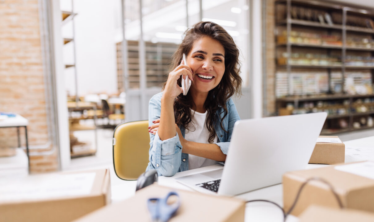 Das Foto zeigt eine weibliche Unternehmerin die vor einem Laptop sitzt und telefoniert, und Pläne für ihr E-Commerce-Business macht.